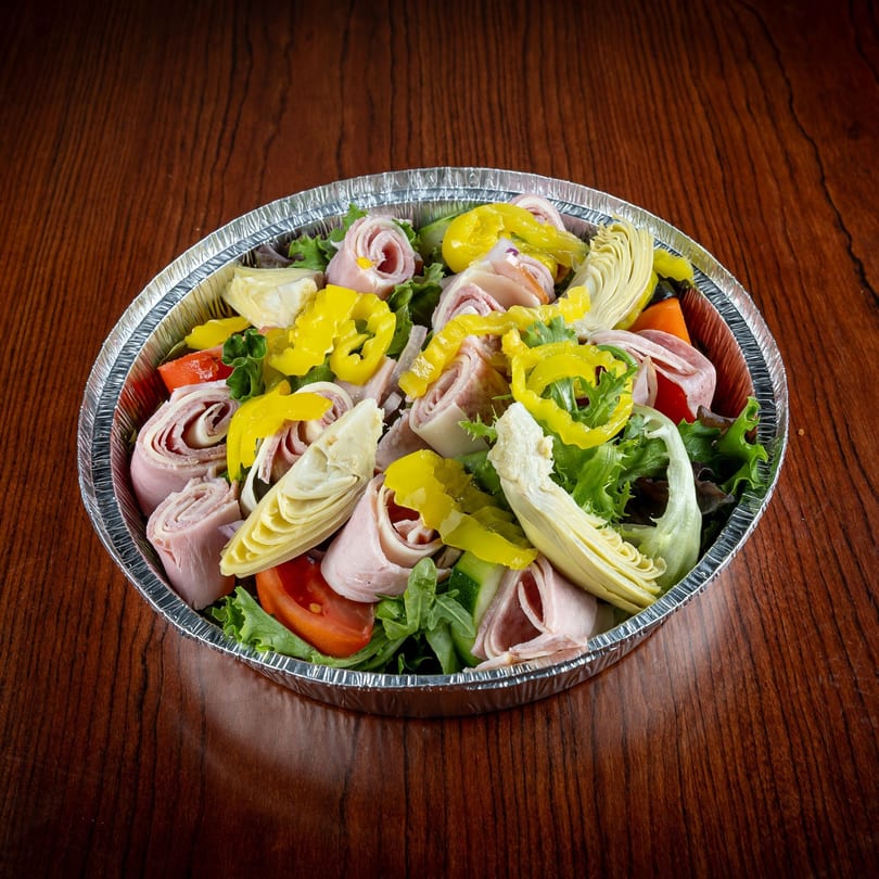Antipasto salad with rolled meats, artichoke hearts, pepperoncini, and lettuce in a foil container.